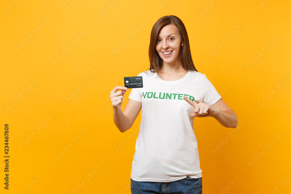 Portrait of young woman in white t-shirt with written inscription green title volunteer hold credit bank card isolated on yellow background. Voluntary free assistance help, charity grace work concept. Portrait of young woman in white t-shirt with written inscription green title volunteer hold credit bank card isolated on yellow background. Voluntary free assistance help, charity grace work concept.