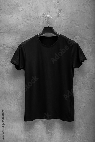 Front side of male black cotton t-shirt on a hanger and a concrete wall in the background. T-shirt without print