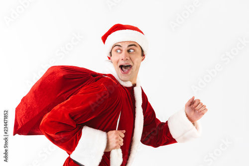 Portrait of positive man 30s in santa claus costume and red hat running with gift bag over shoulder, isolated on white background in studio