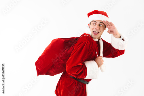 Portrait of optimistic man 30s in santa claus costume and red hat running with gift bag over shoulder, isolated on white background in studio