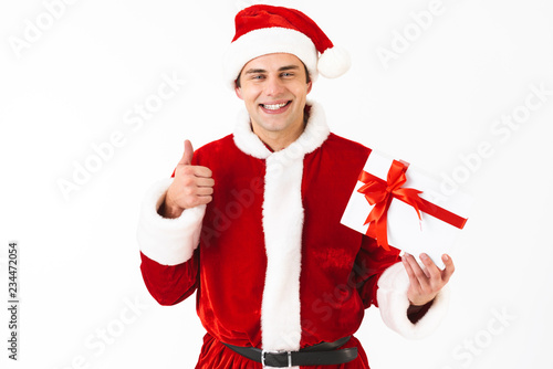 Portrait of cheerful man 30s in santa claus costume and red hat holding present box, isolated on white background in studio