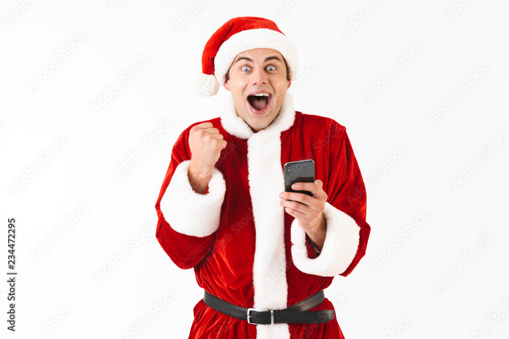 Image of excited man 30s in santa claus costume holding mobile phone, isolated on white background in studio Image of excited man 30s in santa claus costume holding mobile phone, isolated on white background in studio