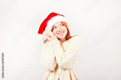Close up portrait of beautiful redheaded young woman wearing Santa Claus hat   white sweater with satisfied facial expression. Cute female celebrating New Year. Copy space  isolated white background.