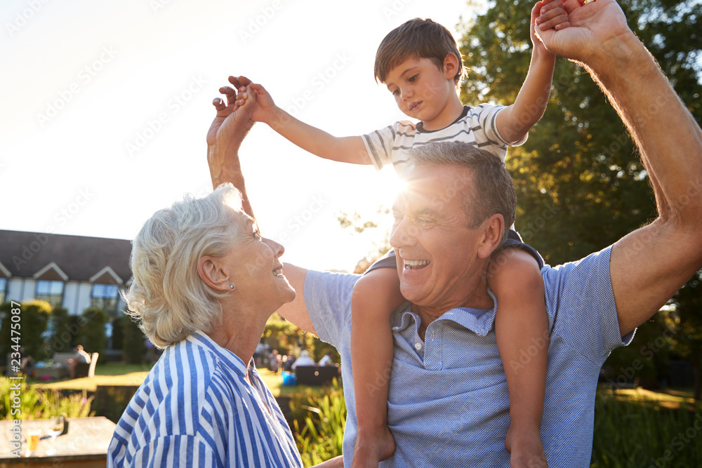 Fototapeta premium Grandparents Giving Grandson Ride On Shoulders In Summer Park Against Flaring Sun