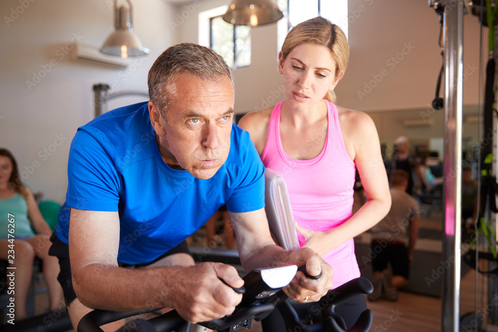 Senior Man Exercising On Cycling Machine Being Encouraged By Female Personal Trainer In Gym Senior Man Exercising On Cycling Machine Being Encouraged By Female Personal Trainer In Gym