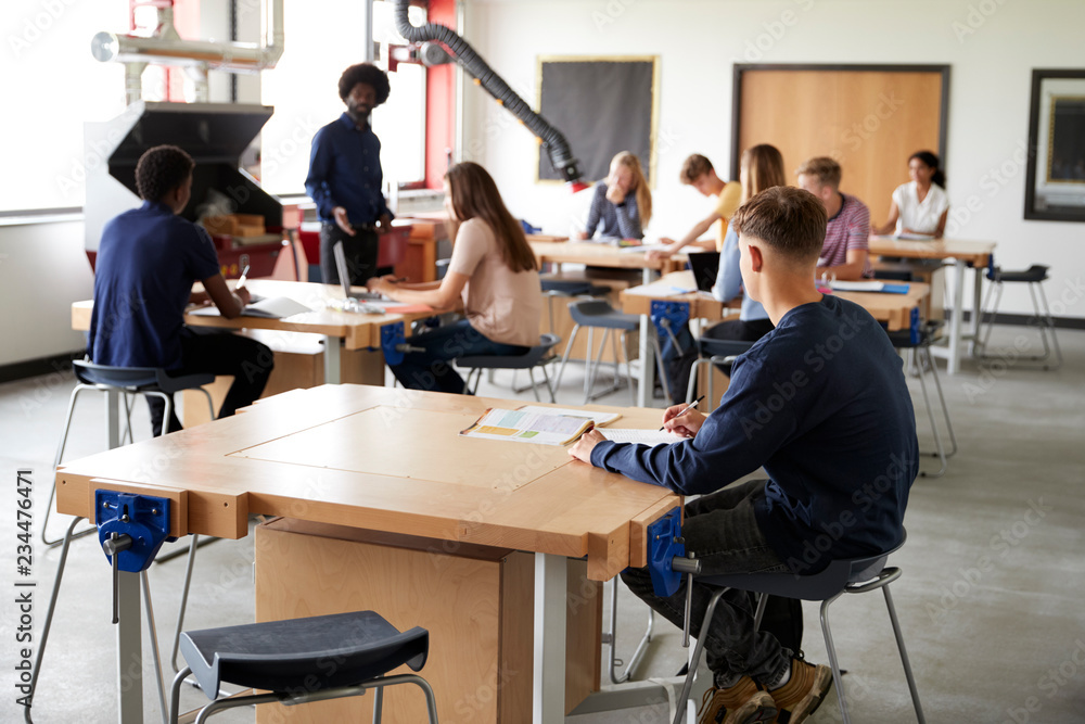 Class Of High School Students Sitting At Work Benches Listening To Teacher In Design And Technology Lesson Class Of High School Students Sitting At Work Benches Listening To Teacher In Design And Technology Lesson