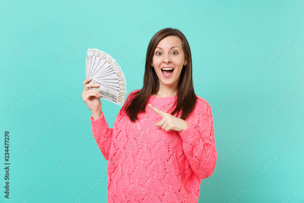 Excited young woman in knitted pink sweater pointing index finger on lots bunch of dollars banknotes, cash money in hand isolated on blue wall background. People lifestyle concept. Mock up copy space. Excited young woman in knitted pink sweater pointing index finger on lots bunch of dollars banknotes, cash money in hand isolated on blue wall background. People lifestyle concept. Mock up copy space.