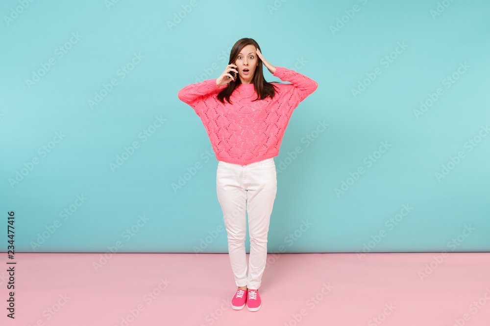 Full length portrait of woman in knitted rose sweater, white pants talking on mobile phone isolated on bright pink blue pastel wall background in studio. Fashion lifestyle concept. Mock up copy space.