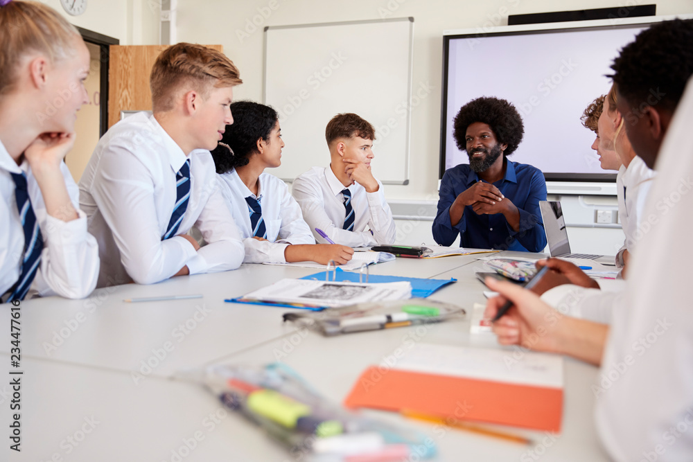 Male High School Teacher Sitting At Table With Teenage Pupils Wearing Uniform Teaching Lesson Male High School Teacher Sitting At Table With Teenage Pupils Wearing Uniform Teaching Lesson