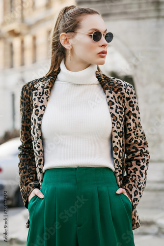 Outdoor fashion portrait of young beautiful confident woman wearing trendy leopard print blazer, white turtleneck, green high-waisted trousers, stylish sunglasses, posing in street of european city