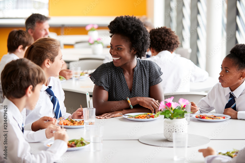 Female Teacher With Group Of High School Students Wearing Uniform Sitting Around Table And Eating Lunch In Cafeteria Female Teacher With Group Of High School Students Wearing Uniform Sitting Around Table And Eating Lunch In Cafeteria