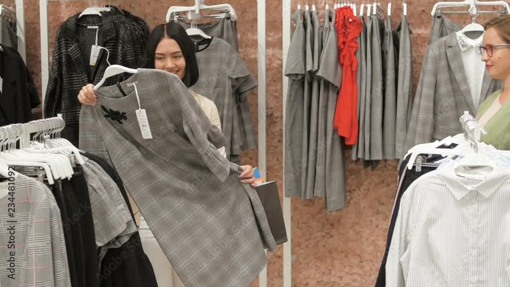 Two young women walking through the mall. Woman shopping. Young woman chooses a sweather in a clothing store and consults with friend. Trying on dresses Two young women walking through the mall. Woman shopping. Young woman chooses a sweather in a clothing store and consults with friend. Trying on dresses
