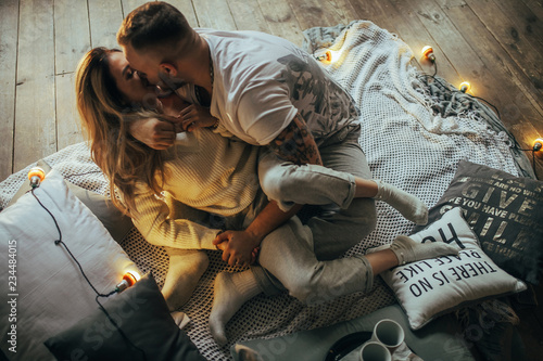 Young couple kissing on background of wooden floor, coverlet, pillows and glowing lightbulbs.