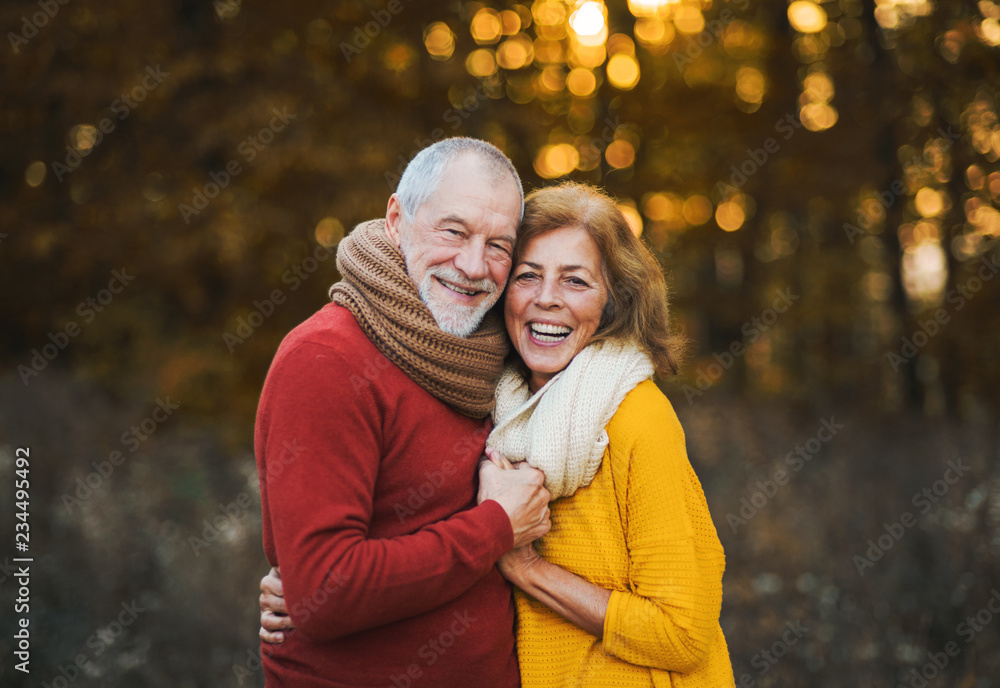 A senior couple standing in an autumn nature at sunset, hugging.
