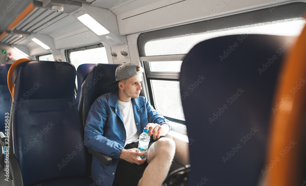 Young man in a cap and a bottle of water in his hands travels by train and looks in the window. Student goes on a train. Young man in a cap and a bottle of water in his hands travels by train and looks in the window. Student goes on a train.