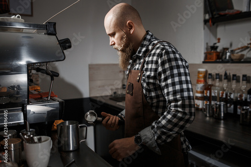 Brutal young barista in an apron makes coffee at the bar in a modern cafe