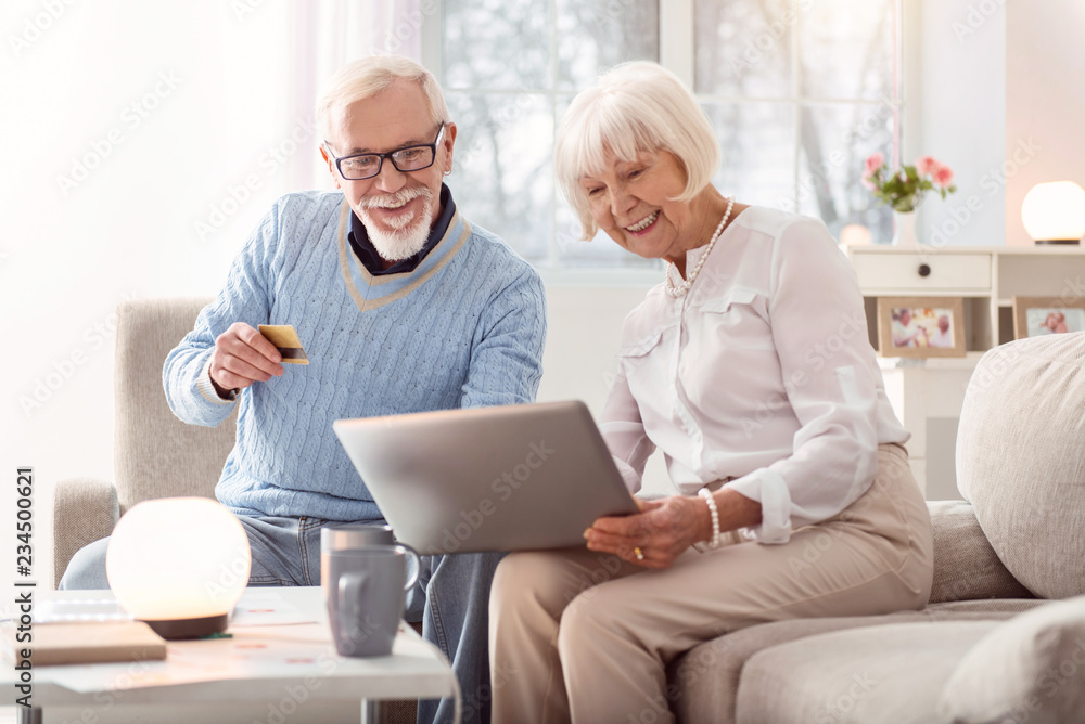 Sincere smiles. Elderly man and woman sitting on the sofa and smiling to each other while looking through the pictures in internet