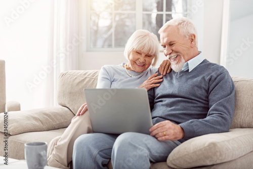 Have a look. Grey-haired cheerful woman sitting on the sofa while showing beautiful pictures to her husband