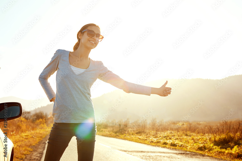 attractive smiling woman in sunglasses is standing on the road near car with far mountains background and she is stopping cars. hitchhiking and car traveling concept