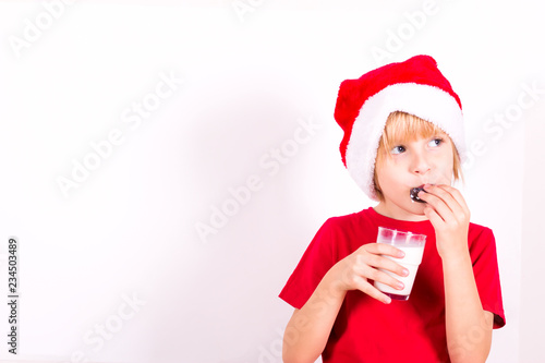 Happy boy in Santa red hat holding milk and chocolate chip cookies in hand. Christmas concept.