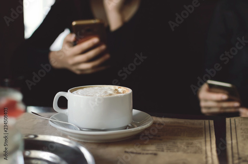 A cup of cappuccino coffee on a table in a cafe. People drink coffee in a cafe and use mobile phones. Close-up, selective focus.