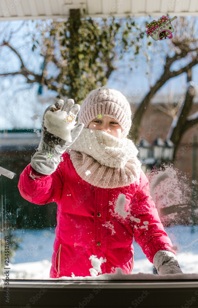 Seven-year-old cute girl in winter clothes is standing outside the door, on  the street with snow in her hands and looking at the house smiling. Stock  Photo