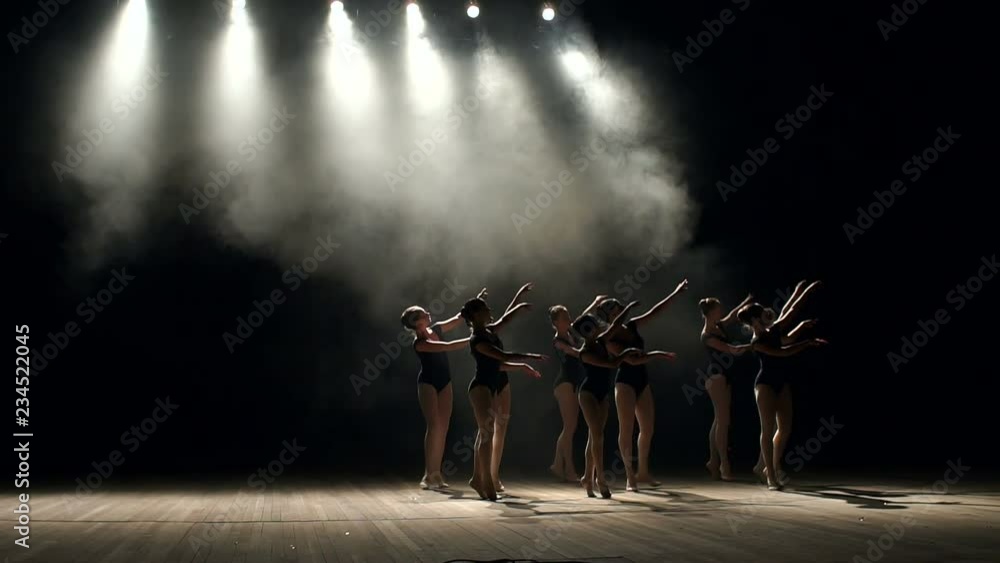 Young teen girls perform ballet on stage in smoke on black background. A choreographed dance of a group of graceful pretty young ballerinas practicing on stage in a classical ballet school.