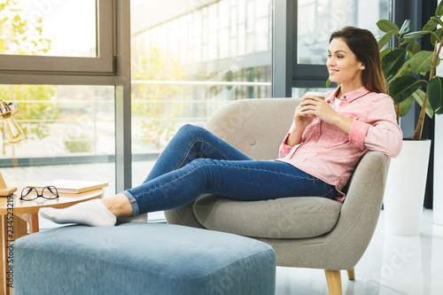 Young smiling woman sitting on sofa while drinking hot coffee. Young brunette woman thinking at home in a leisure time. Happy girl relaxing at home on big chair.