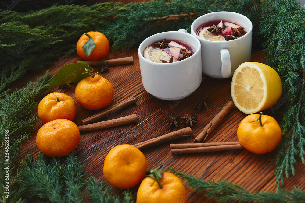 Christmas hot mulled wine in two small white cups with spices and citrus fruits on a wooden background and coniferous branches, top view