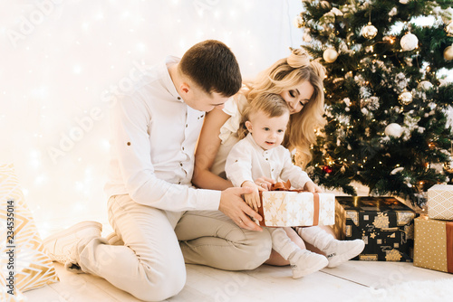 Portrait of beautiful young family on Christmas tree and white cotton background. Attractive parents and a little son open new year gifts © ksyusha_yanovich