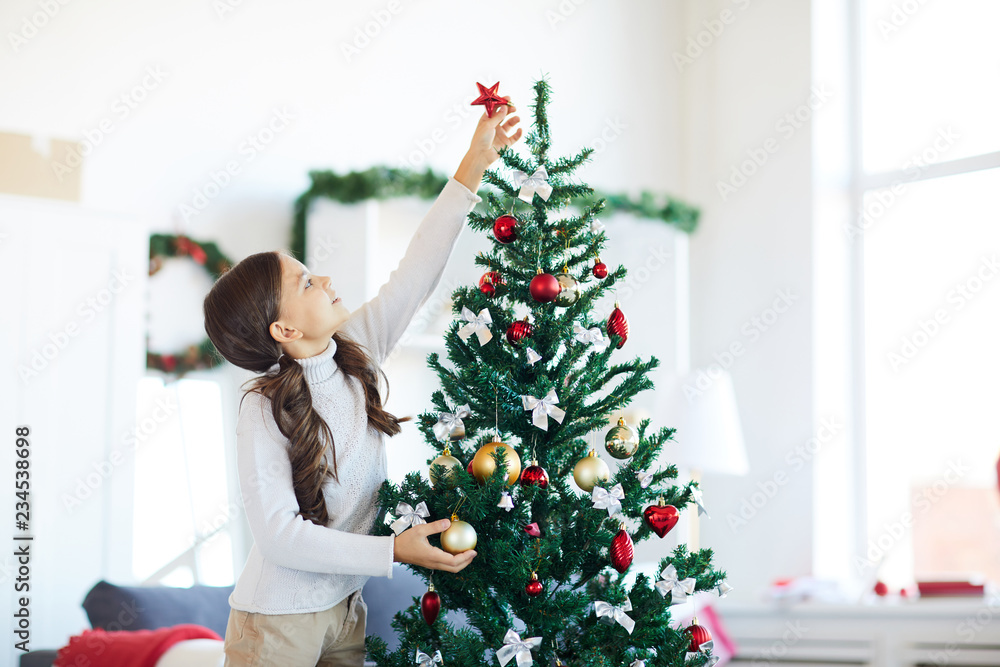 Little girl in white jumper reaching for top of xmas tree to put red star Little girl in white jumper reaching for top of xmas tree to put red star
