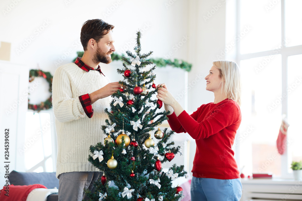 Young casual couple standing by xmas tree and decorating it on holiday eve Young casual couple standing by xmas tree and decorating it on holiday eve