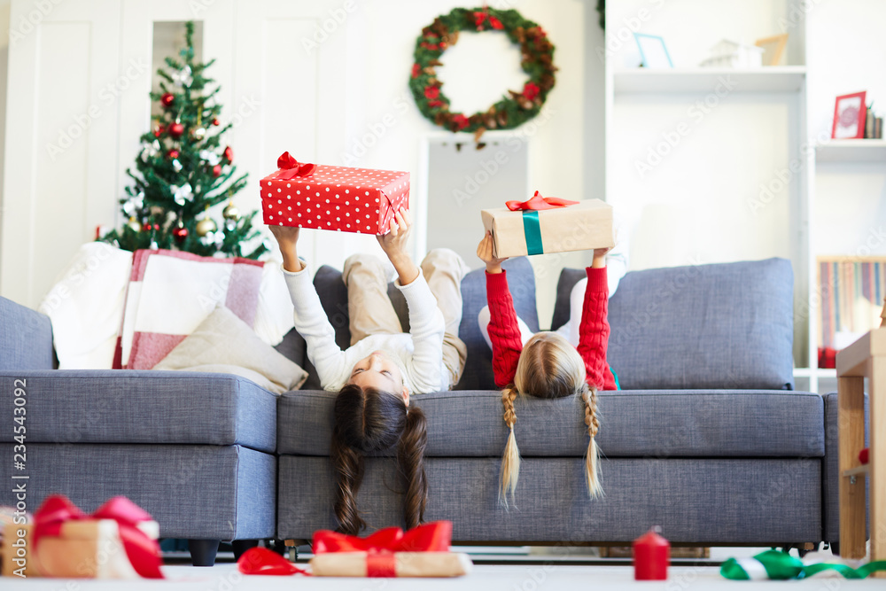 Fototapeta premium Two little girls lying on sofa and holding giftboxes while stretching arms in front of themselves