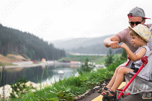 The happy young father sitting on the road near a lake near his little son in a stroller and they beating by fists