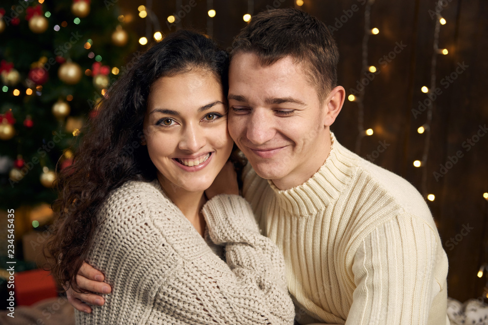 young couple is in christmas lights and decoration, dressed in white, fir tree on dark wooden background, winter holiday concept young couple is in christmas lights and decoration, dressed in white, fir tree on dark wooden background, winter holiday concept