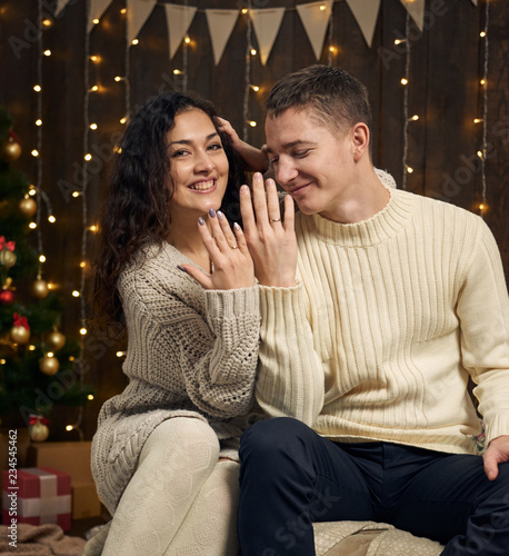 young couple is in christmas lights and decoration, dressed in white, fir tree on dark wooden background, winter holiday concept