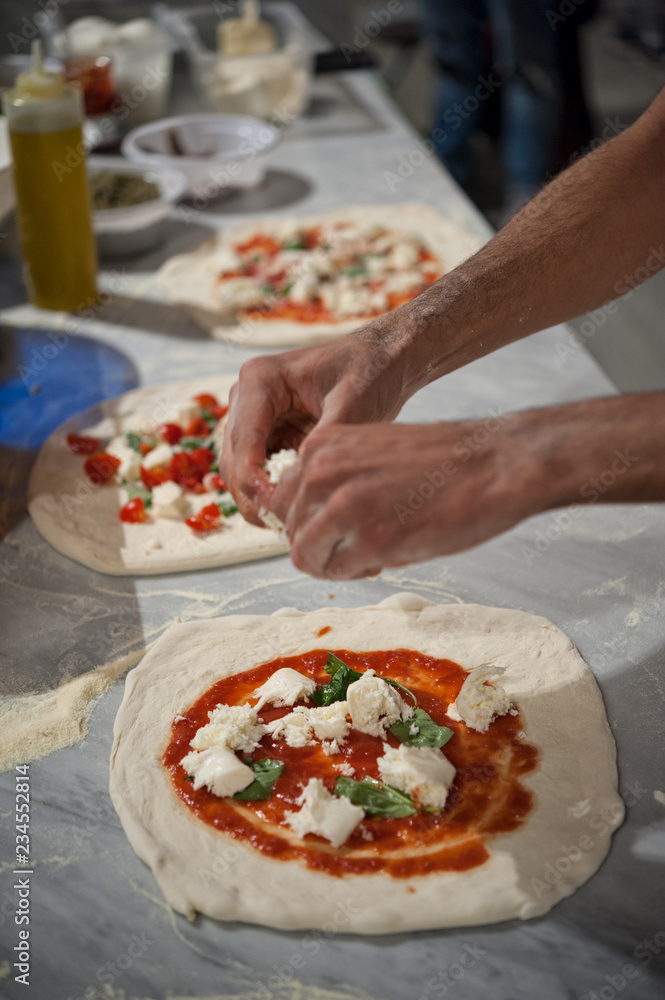 Preparing Pizza Margherita on a marble countertops. Pizzaiolo puts pieces of mozzarella and basil leaves over a raw pizza dough with tomato sauce. Selective focus   