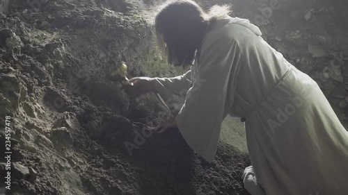 A man like Jesus Christ is digging a hole in the dungeon, a man is searching for a precious treasure, a man in a white robe is digging the earth with a stick and finds gold, Jesus hides the holy grail photo