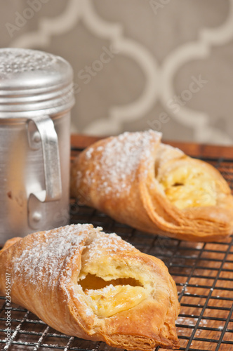 Front view, close up of two freshly baked cheese danish pasteries with powdered shgar and a sugar sprinkler, cooling on a metal, wire rack on a wood cutting board