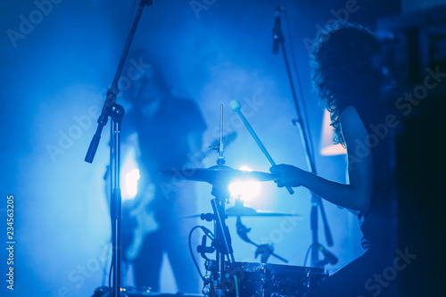  Female drummer with long curly hair in front of singing guitarist on a stage in a bright blue lights playing rock show