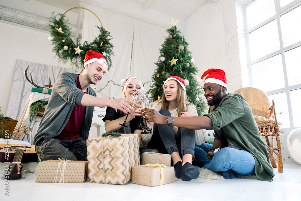 Fototapeta premium Young African man and Caucasian man and women, 20 to 30 years old, with Santa hats clink glasses drinking champagne for christmas or xmas and new year celebration party.