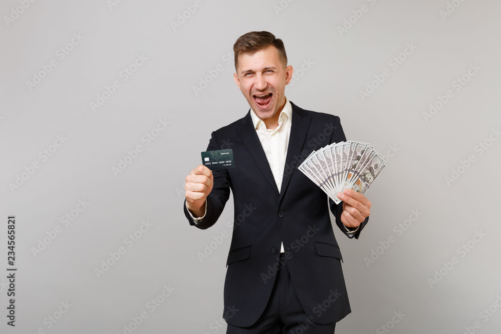 Happy young business man in suit screaming hold credit bank card lots bunch of dollars banknotes cash money isolated on grey background. Achievement career wealth business concept. Mock up copy space. Happy young business man in suit screaming hold credit bank card lots bunch of dollars banknotes cash money isolated on grey background. Achievement career wealth business concept. Mock up copy space.