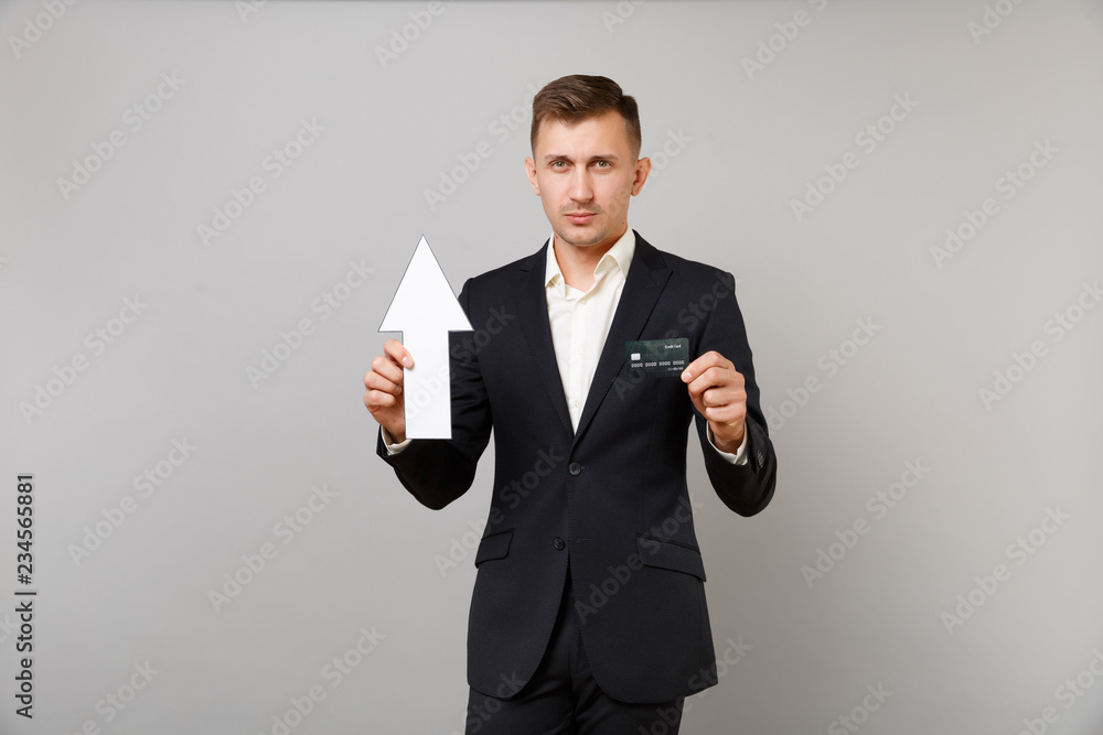 Handsome young business man in classic black suit, shirt holding up arrow, credit bank card isolated on grey wall background in studio. Achievement career wealth business concept. Mock up copy space. Handsome young business man in classic black suit, shirt holding up arrow, credit bank card isolated on grey wall background in studio. Achievement career wealth business concept. Mock up copy space.