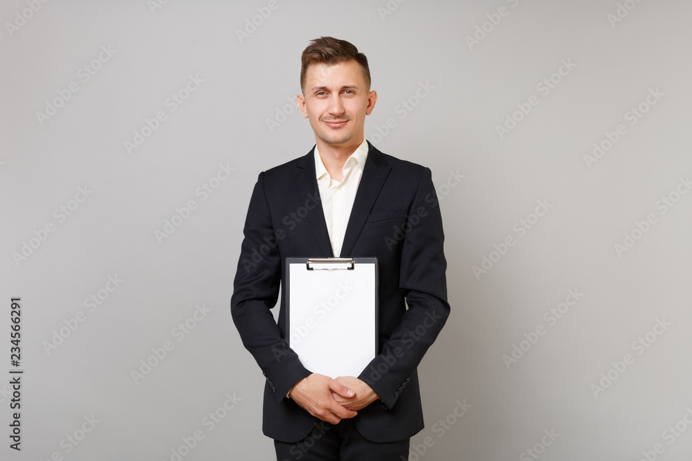 Handsome young business man in classic black suit, shirt holding clipboard with blank empty sheet workspace isolated on grey background. Achievement career wealth business concept. Mock up copy space. Handsome young business man in classic black suit, shirt holding clipboard with blank empty sheet workspace isolated on grey background. Achievement career wealth business concept. Mock up copy space.