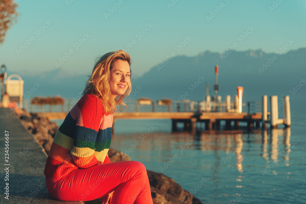 Beautiful woman resting by the lake at sunset on warm evening, wearing colorful pullover
