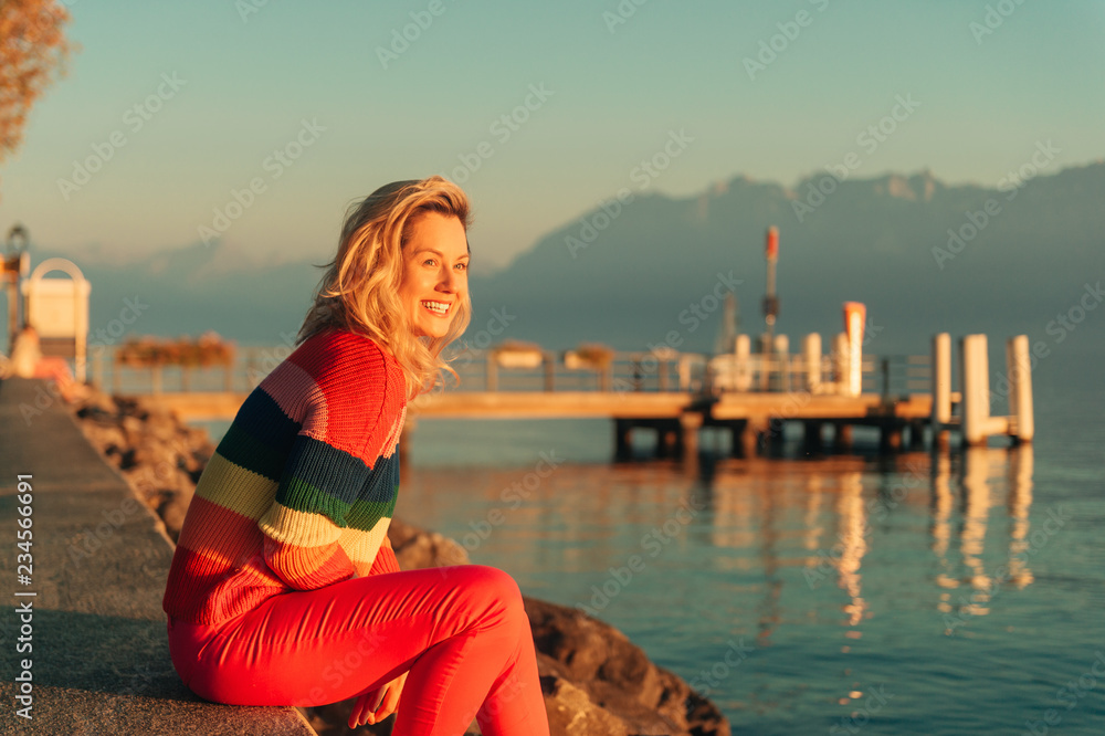 Beautiful woman resting by the lake at sunset on warm evening, wearing colorful pullover