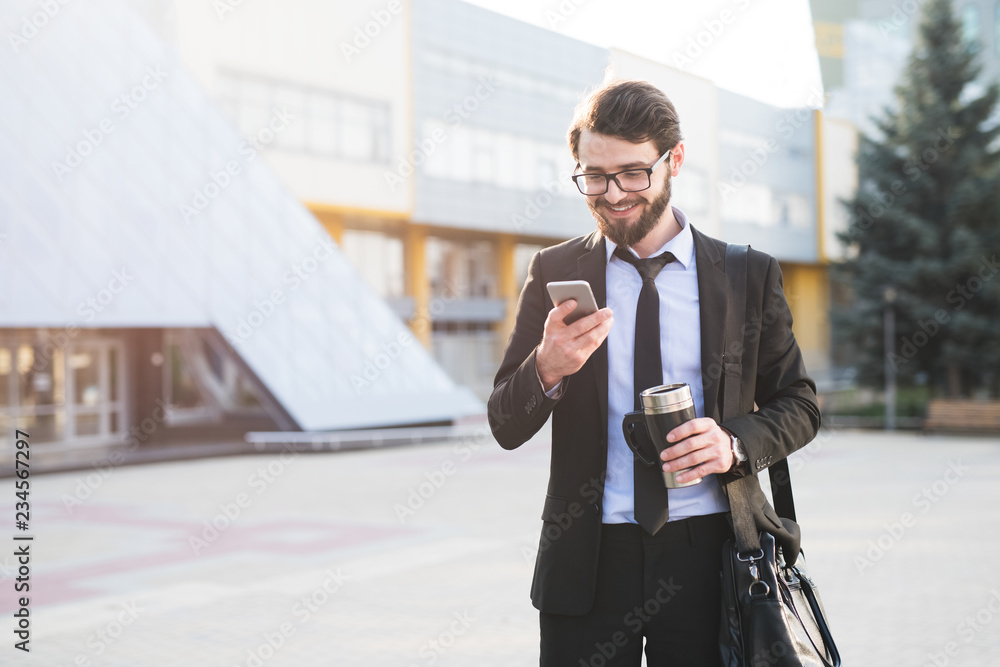 Portrait of smiling businessman using smartphone near office build with coffee thermo mug in hand Portrait of smiling businessman using smartphone near office build with coffee thermo mug in hand