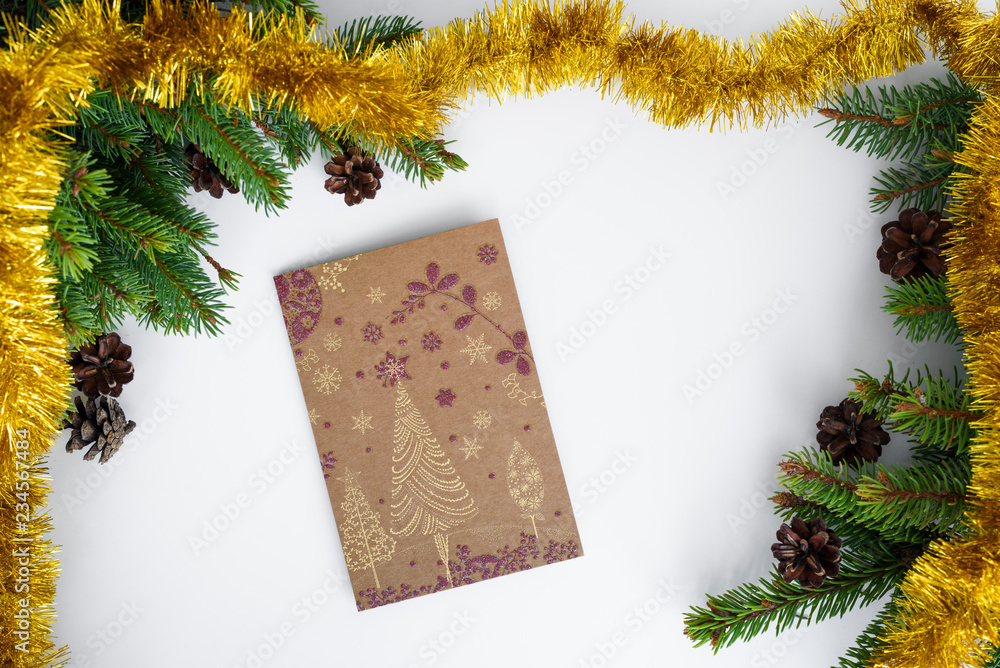 Fototapeta premium Christmas greeting card is lying on white table, surrounded by festive decorations like tree branches, cones and gold tinsel. Merry Christmas.