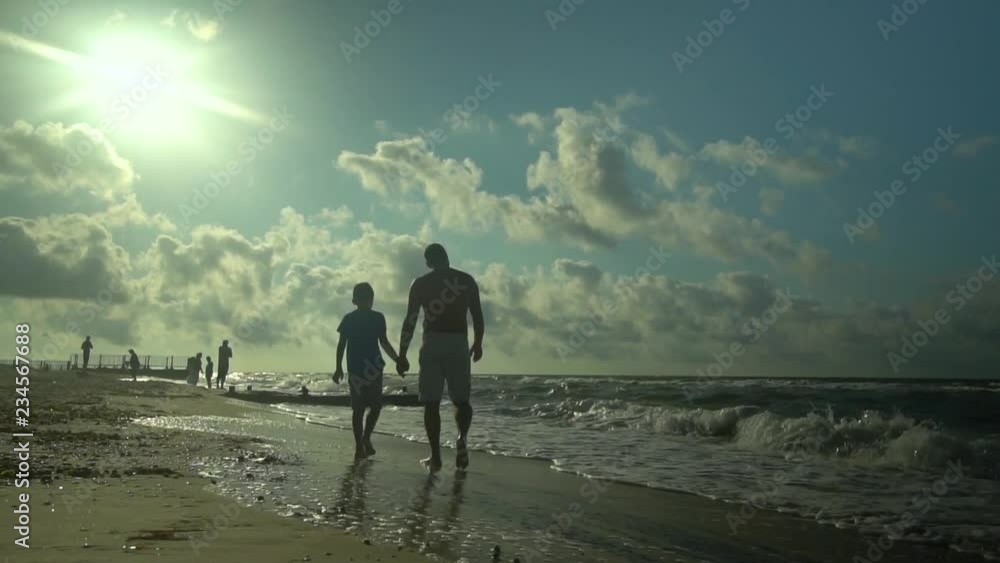 A young father walks with his son along the seashore. The sun shines brightly. They are talking. Family happiness. Trust relationship between father and son. Slow motion. A young father walks with his son along the seashore. The sun shines brightly. They are talking. Family happiness. Trust relationship between father and son. Slow motion.