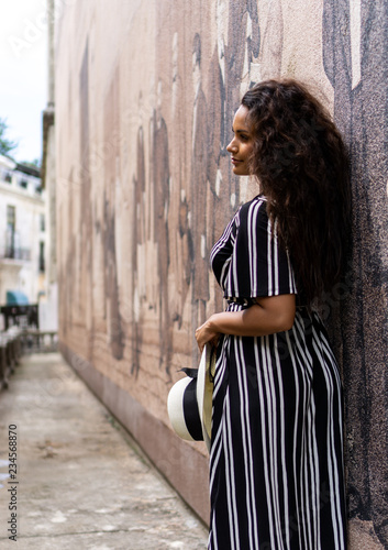 Young lady with curly hair and a black and white dress posing in front of an artistic wall in Cuba Havana holding a white and black hat in her hand. Shot is taken from the side showing her profile. © Saga_bear